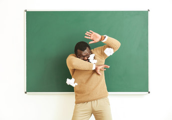Naughty pupils throwing crumpled paper in African-American teacher near blackboard