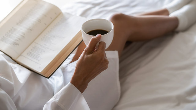Asian woman holding cup of coffee and reading a book on the bed after get up in morning. Morning lifestyle concept.