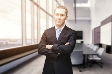Front view of portrait of young Caucasian businessman with arms crossed smiling to camera. He is standing in the glass walled lobby of a modern business building. Modern corporate