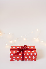 Gift box in red paper packaging with white polka dots, tied with a red satin ribbon with a bow and a glass heart on a white background with lights