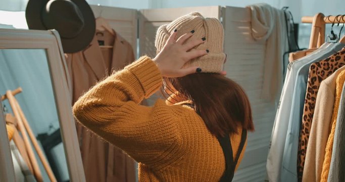 Close up of the beautiful cheerful young female fashion blogger taking on hat in front of the mirror, then turning facce to the camera and smiling to the camera in the designer's studio. Portrait.