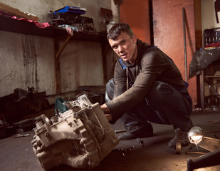 Auto mechanic squatting working on car detail on floor at auto repair shop, looking to the camera. Strong repairman in dirty workwear in action. Tools, spare parts and garage interior on background