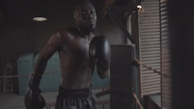 Waist-up handheld shot of young African American boxer with bare torso, in shorts and gloves ferociously punching heavy cylindrical bag in dark rundown fight club