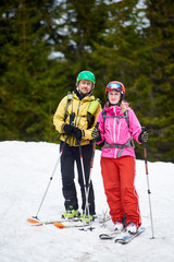 Vertical portrait snapshot of happy couple woman and man skiers posing looking in camera standing together on ski on snow-covered slope with ski poles in hands. Green trees on blurred background