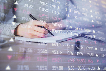 Hands writing information about stock market in notepad. Forex chart holograms in front. Concept of...