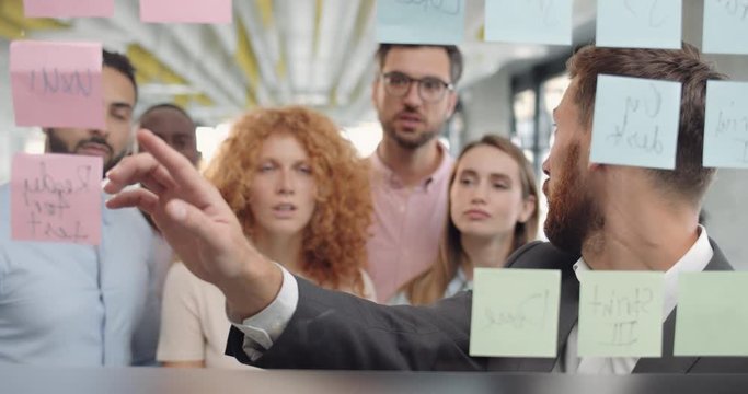 Group of male and female workers having lively discussion near glass board with sticky notes. Male business coach in 30s standing and talking with multiethnic team at corporate training.
