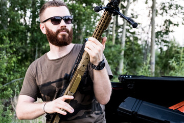 A man with glasses with a beard holds a sniper rifle