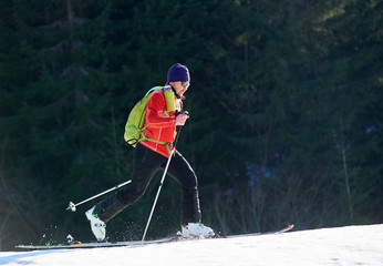 Male skier backpacker in sunglasses walking on skis up snowy hill on background of ever-green...