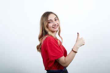 Cheerful young caucasian woman in fashion red shirt staying sideways, looking at camera, showing thumbs at camera isolated on white background in studio. People sincere emotions, lifestyle concept.