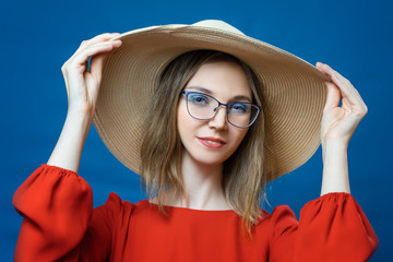 A beautiful young girl in a hat and glasses posing on a blue background in red clothes. Elegant girl model appearance. The concept of beauty in glasses