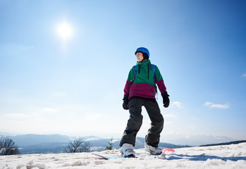 Fototapeta na wymiar Snowboarder young woman in helmet standing on snowboard ready to ride on copy space background of bright blue sky and snowy mountain peaks. Extreme winter sports, active lifestyle concept.