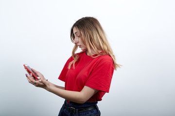 Attractive young caucasian woman in fashion red shirt keeping telephone, looking on it isolated on white background in studio. People sincere emotions, lifestyle concept.