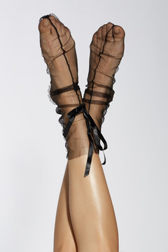 Cropped subject shot of lifted female legs in black nylon transparent socks with middle stripes on the front side, lace in the back and silk strings. The photo is made on the light-gray background. 