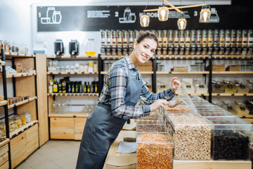 Mixed Race Woman Owner Selling Superfoods in Zero Waste Shop. Lots of Healthy Food in Glass Bottles on Stand in Grocery Store. No plastic Conscious Minimalism Vegan Lifestyle Concept. Selective focus