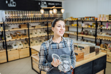 Mixed Race Woman Owner Selling Superfoods in Zero Waste Shop. Lots of Healthy Food in Glass Bottles on Stand in Grocery Store. No plastic Conscious Minimalism Vegan Lifestyle Concept. Selective focus