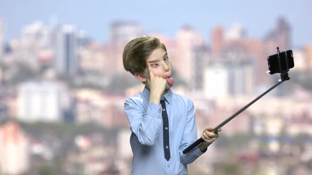 Child boy grimacing while taking selfie. Preteen caucasian boy showing tongue to camera. Blurred city background.