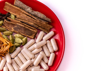Top view macro shot of cinnamon sticks, cardamom, clove and nutmeg aril with herbal capsules in a red plate on copy space white background. Ayurvedia concept