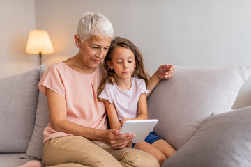 Smiling granddaughter and grandmother using digital tablet on sofa in living room. A grandmother...