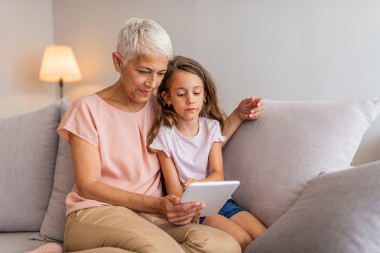 Smiling granddaughter and grandmother using digital tablet on sofa in living room. A grandmother and granddaughter are hugging looking at a digital tablet. Small girl and her grandmother with tablet 