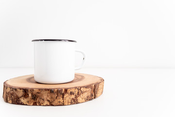 Enamel mug mock up with wooden cut tree section on white table. Boho design of tin cup on white background with copy space for image or text