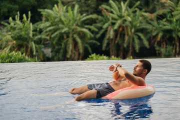 Attractive man floating on inflatable ring in swimming pool and looking at smartchasy or tracker....