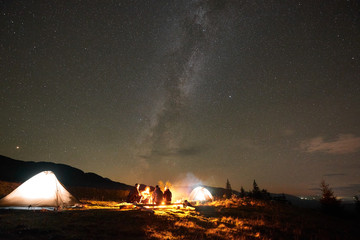 Night camping under stars. Group of four hikers, men and woman with guitar sitting by burning campfire at illuminated tourist tents under summer night starry sky and Milky Way constellation.