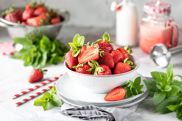 Fresh strawberries in a bowl and sprigs of mint on a white concrete table. Ingredient for smoothies. Bottle of milk, red glass mug and a blender. The recipe for the holiday Valentine's Day. close up