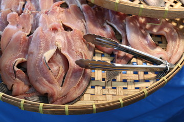 Sun dried striped snake-head fishes are on rice winnowing basket and stainless tongs, Thailand.