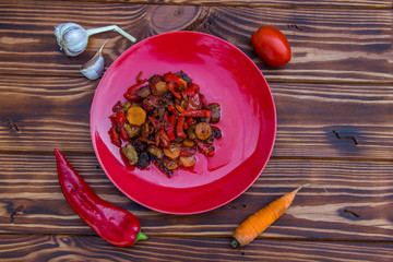 fried vegetables garlic tomato carrot onion peppers with meat in a red plate on a brown wooden background