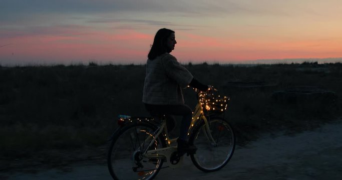 Silhouette di una ragazza va in giro in bicicletta di notte sulla spiaggia. Striscia illuminata di luci led.