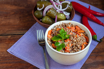 Buckwheat porridge in a bowl with stewed carrots. In the background are fermented pickled cucumbers and red chili peppers.