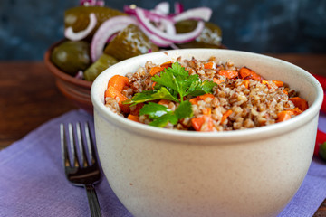 Buckwheat porridge in a bowl with stewed carrots. In the background are fermented pickled cucumbers and red chili peppers.