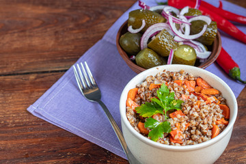 Buckwheat porridge in a bowl with stewed carrots. In the background are fermented pickled cucumbers and red chili peppers.