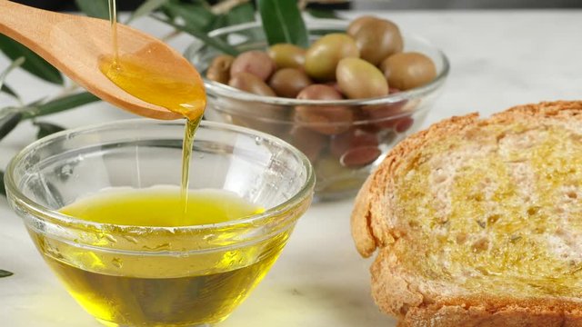 extra virgin oil, genuine Italian ingredient falls by tablespoon on glass bowl. Bread and olives on background. Healthy food, natural product.