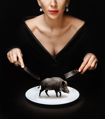 A girl with a knife and fork in her hands in a black dress is holding a white plate in which to be a wild boar on a black table