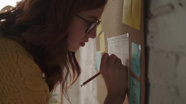 Young modern woman goes to wall with cork board, writes on stickers