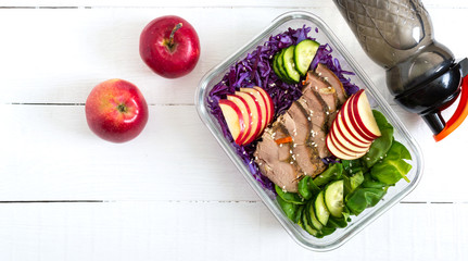 Tasty healthy lunch of vegetables, baked turkey and a bottle of water. Salad of red cabbage, spinach, apples, fresh cucumbers with diet meat in a glass lunchbox on a white wooden background.