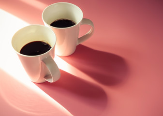 Two cups of black coffee on pink background with morning sunlight.