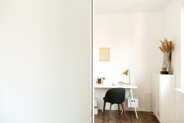 Modern Scandinavian interior design concept. Home office workspace cabinet. White stylish freelancer working room. Artist studio with table, chair, chest of drawers, lamp, flowers, wooden floor.