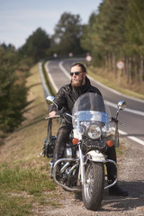Fototapeta na wymiar Handsome bearded biker with long hair in black leather jacket and sunglasses sitting on modern motorcycle on roadside, on blurred background of empty twisty road with white marking on sunny summer day