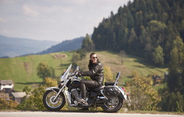 Side view of handsome bearded biker in black leather jacket and sunglasses sitting on modern motorcycle on roadside on blurred background of foggy spruce forest, green hills and bright sky.