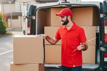 Image of smiling delivery man writing while standing with parcel boxes