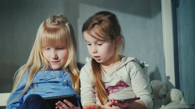 Two girls 5 and 7 years are vividly discussing something, they use their smartphones. Free time of modern children concept
