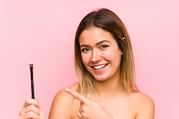 Obraz premium Young caucasian woman holding a eyebrush isolated smiling cheerfully pointing with forefinger away.