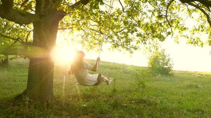 young girl swinging on swing on an oak branch in sun. Dreams of flying. Happy childhood concept. Beautiful girl in white dress in park. teen girl enjoys flight on swing on a summer evening in forest - obrazy, fototapety, plakaty