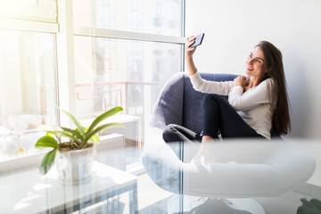 Young beautiful woman making selfie photo using cellphone while sitting in chair. Pretty lady shooting herself for blog or social network post during vacation