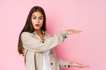 Young caucasian woman posing in a pink background shocked and amazed holding a copy space between hands.