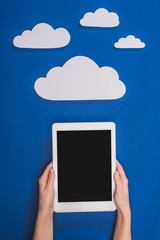 cropped view of woman holding digital tablet near white paper cut clouds on blue background