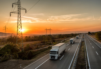 Fototapeta premium Convoy of White transportation trucks in line as a caravan or convoy on a country highway under an amazing sunset sky