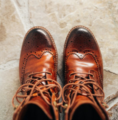 A pair of Brown natural leather medium boots with unlaced shoelaces standing on natural stone floor top view.